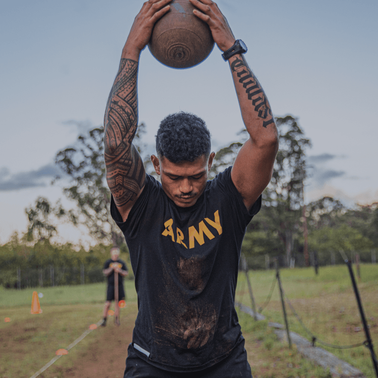 Male soldier lifts medicine ball overhead in ACFT Power Throw event, tattoo visible.