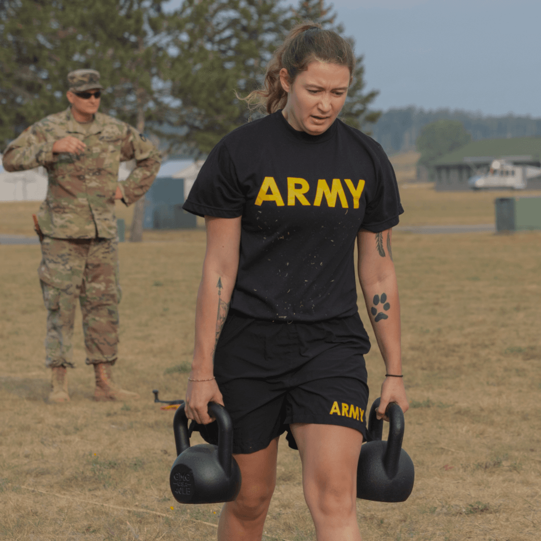 Focused female soldier carries kettlebells in the Sprint-Drag-Carry with a grader watching.